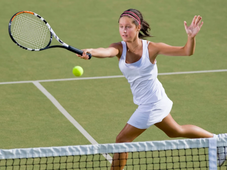 A female tennis player in white outfit dives to hit a volley near the net on a grass court, racquet extended and ball approaching.