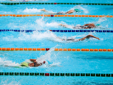 Several swimmers race in a blue pool, each in their own lane marked by orange, green, and blue lane lines, splashing as they swim freestyle.