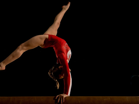 A gymnast in a red leotard balances on a beam, performing a back handspring with one hand on the bar and legs extended in a split arc.