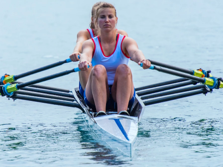 A female rower in a single scull is sprinting on calm water, gripping the oars with focused effort. Always ending the sentence.