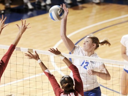 A volleyball player in white jersey (#12) spikes over a net while two red-jersey blockers jump to block, with another white-jersey player in back.