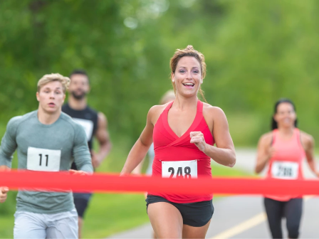 A woman in a red tank top wins a race, crossing the finish line with a smile; two runners close behind on a green path.