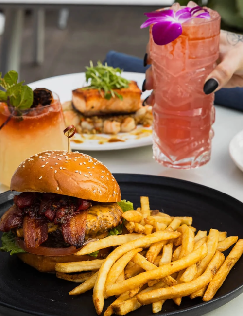 A plated burger with fries, a tall pink cocktail, another dish in the background, and a leafy salad on the side at a table.