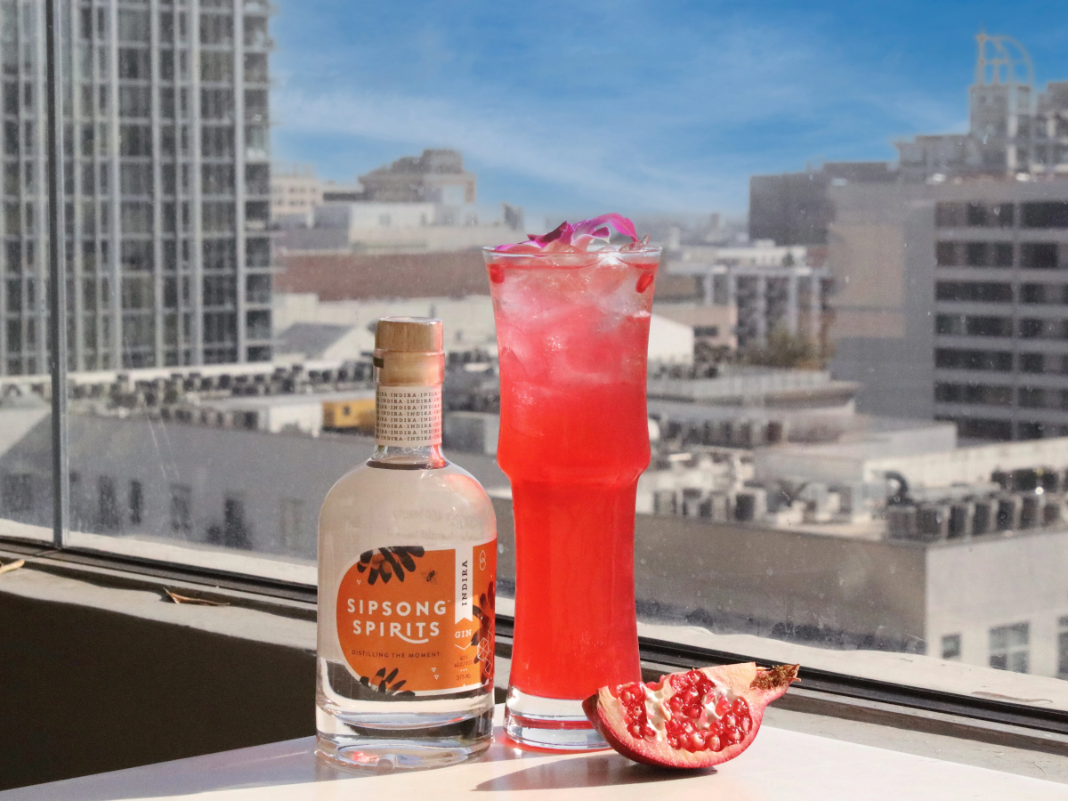 A small bottle of hibiscus syrup beside a tall pink cocktail with ice and a grapefruit garnish on a sunny windowsill, urban skyline in the background.