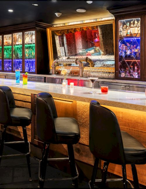 A tidy bar with black stools, a wooden counter, neon-lit shelves, and colorful bottles behind the glass, reminiscent of a cozy lounge bar.