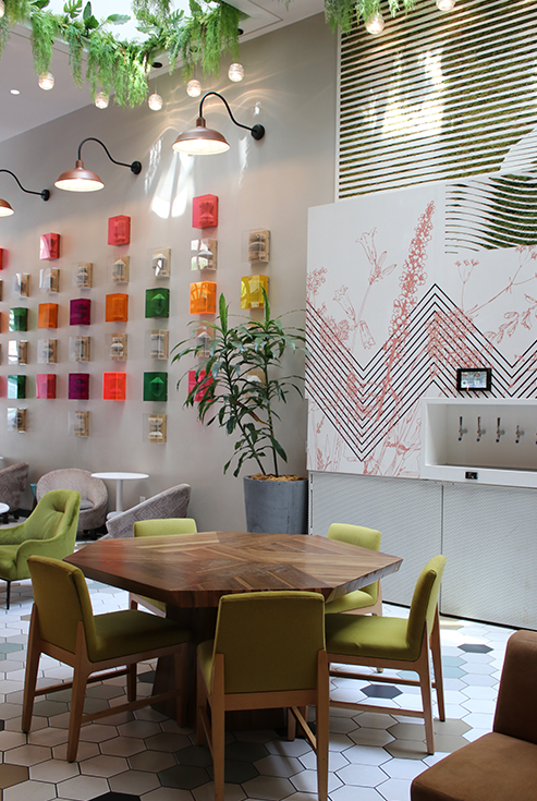 Colorful wall shelves with small colored boxes, round wooden table, green and yellow chairs, indoor plants, modern pendant lights, and large geometric art on a light tiled cafe.