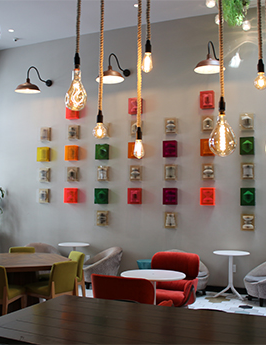 A bright, stylish cafe with colorful cube wall art, hanging Edison bulbs, and a mix of red, yellow, and green chairs around wooden tables. Yes.