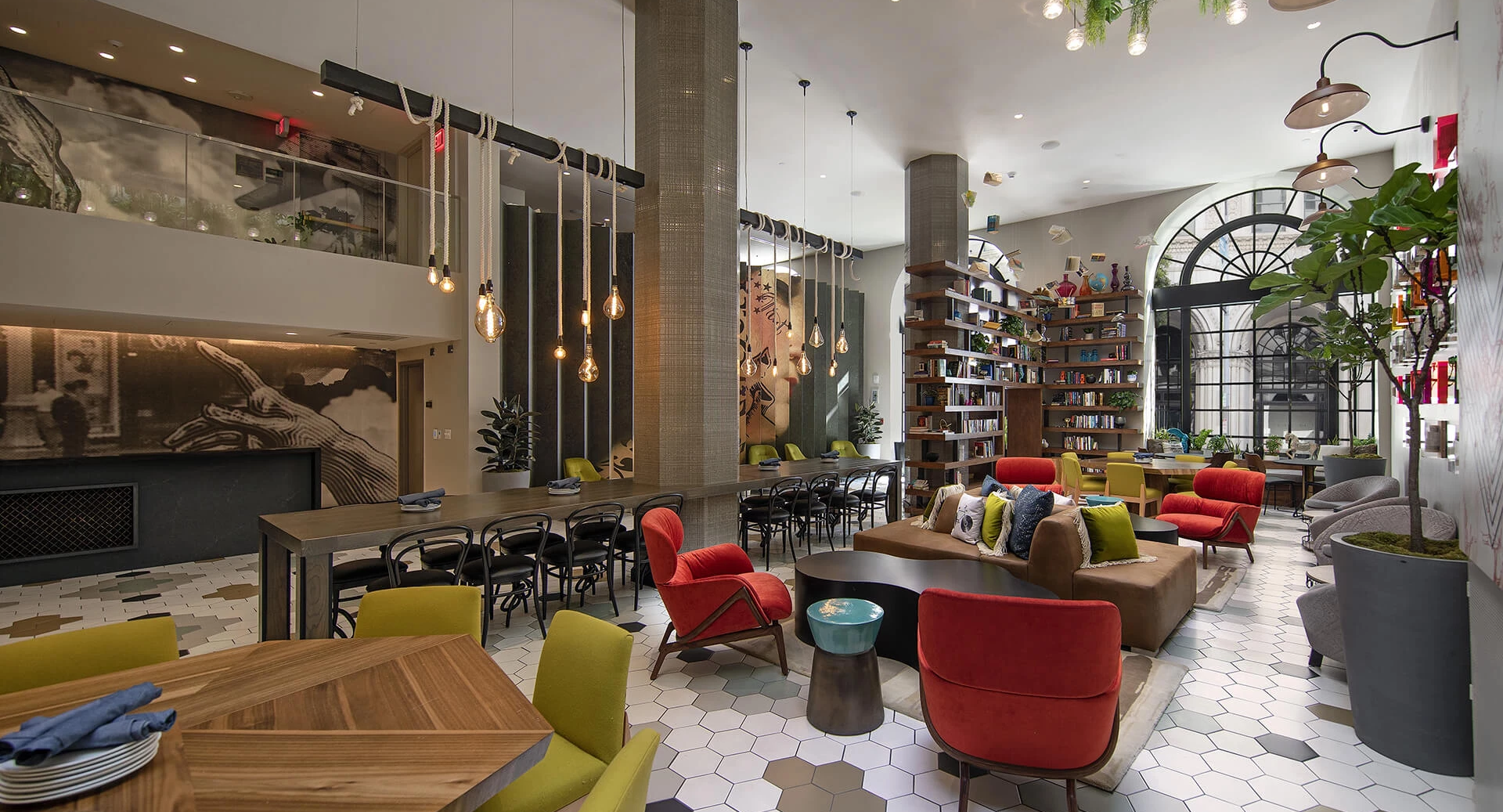 A bright, stylish caf&eacute; with colorful chairs, a long dining table, cozy red sofas, hanging lights, plants, and a wide arched window in the back.