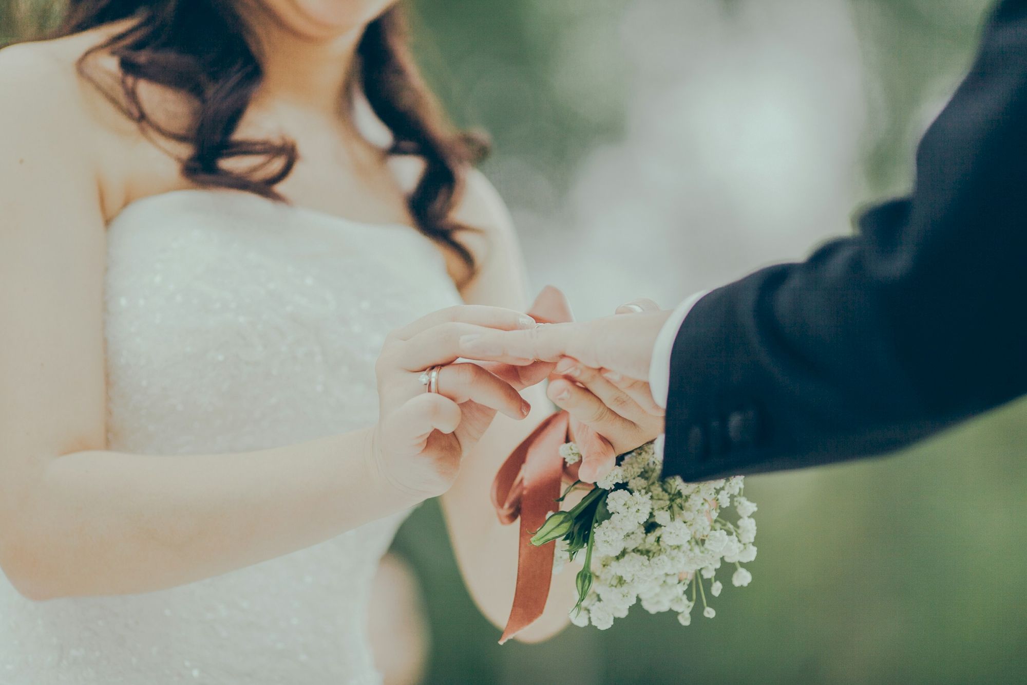 A close-up of a bride and groom exchanging rings; the groom places a ring on the bride&rsquo;s finger, bouquet visible, soft outdoor backdrop.