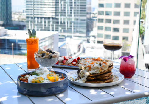 A sunny rooftop feast: skillet shakshuka, baked eggs, fluffy pancakes, fruit-topped cocktails, a carrot shooter, and a glass of red wine.
