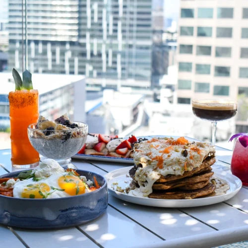 A sunny rooftop feast: skillet shakshuka, baked eggs, fluffy pancakes, fruit-topped cocktails, a carrot shooter, and a glass of red wine.