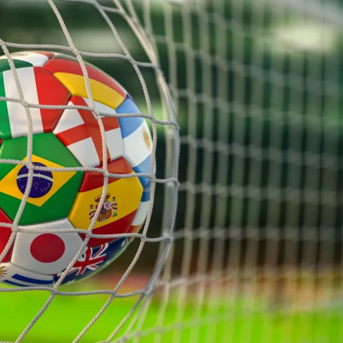 A colorful soccer ball with flags from Portugal, Brazil, Japan, and more, caught in a goal net against a green field, symbolizing international football.