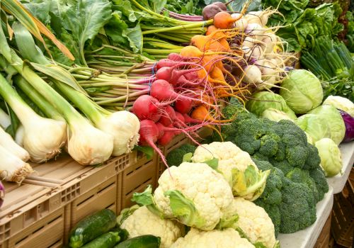 Fresh vegetables on display: white onions, green onions, carrots, radishes, cabbages, broccoli, cauliflower, peppers, and leafy greens arranged in crates and baskets.