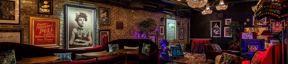 A cozy, eclectic lounge with vintage portraits, plush seating, chandeliers, colorful ottomans, and warm, moody lighting for lounging and socializing.
