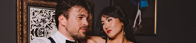 A man in suspenders and a white shirt sits on a leather couch with a woman in a black dress, both holding drinks in a stylish, art-decorated lounge.