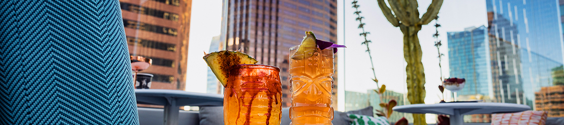 Two tropical cocktails on a wooden tray sit on a rooftop lounge with colorful seating, cactus plants, and city skyscrapers in the background.