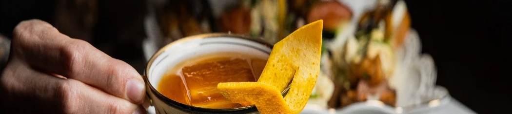 A hand holds a delicate teacup with orange peel garnish, set on a saucer, with a tiered pastries display in the blurred background.