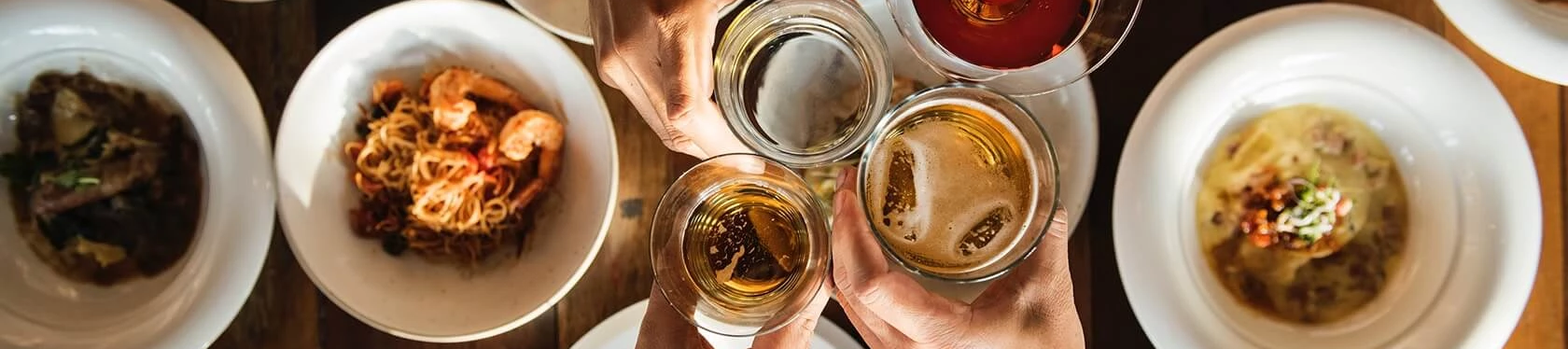 Friends cheers around a table of bowls and plates, clinking drinks in a festive overhead toast ending with a promise of more bites to come.