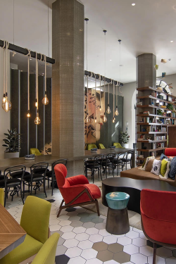 A bright, stylish cafe with modern seating: red armchairs, lime-green chairs, wooden tables, a long bar-height counter, hexagonal tiled floor, shelves, plants, and large arched windows.