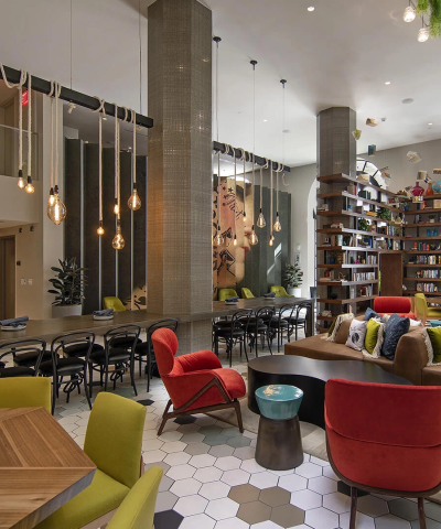A bright, stylish cafe with modern seating: red armchairs, lime-green chairs, wooden tables, a long bar-height counter, hexagonal tiled floor, shelves, plants, and large arched windows.