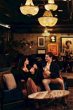 A cozy, dimly lit lounge with chandeliers, vintage decor, and a mix of people chatting on sofas and chairs in a warm, intimate setting.