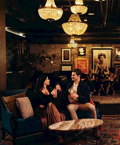 A cozy, dimly lit lounge with chandeliers, vintage decor, and a mix of people chatting on sofas and chairs in a warm, intimate setting.