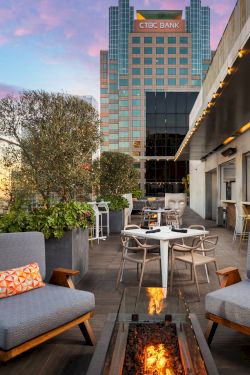 A stylish rooftop lounge with modern seating, a fire pit, city skyline, and a bar area at sunset, inviting relaxation above the streets.