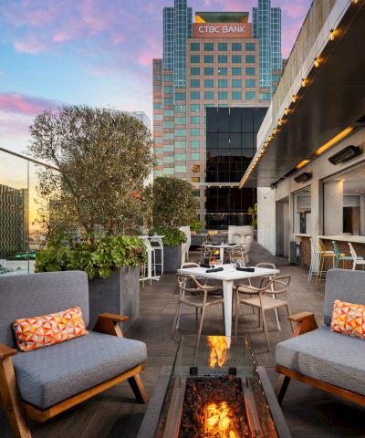A stylish rooftop lounge with modern seating, a fire pit, city skyline, and a bar area at sunset, inviting relaxation above the streets.