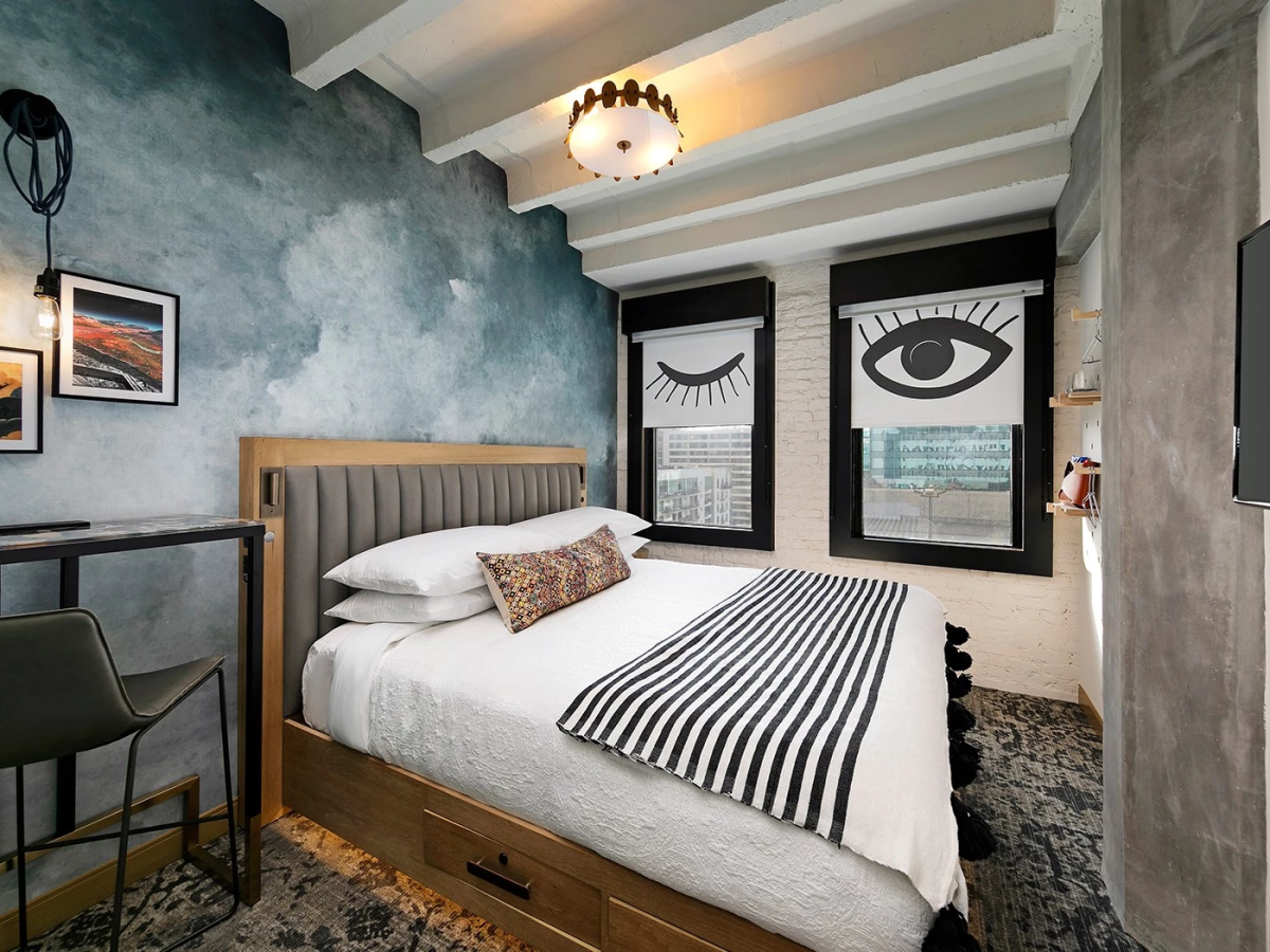 A cozy, stylish bedroom with a tufted headboard, striped bedding, wall art, and large window panels featuring bold eye artwork&mdash;bright and eclectic.