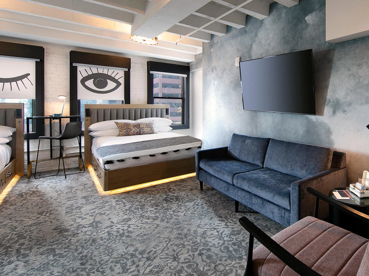 A modern hotel suite with two beds, bold eye artwork, a blue sofa, a wall-mounted TV, and a cozy seating area, all onderlaid by a patterned carpet.