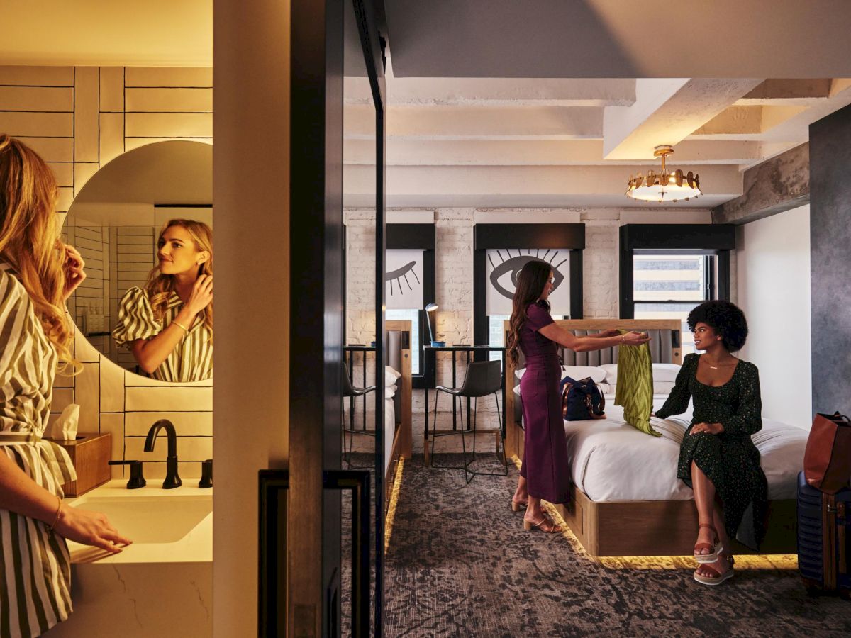 Two women in a hotel suite prep area: one at a sink applies makeup in a mirror, while another woman sits on a bed with a chair nearby.
