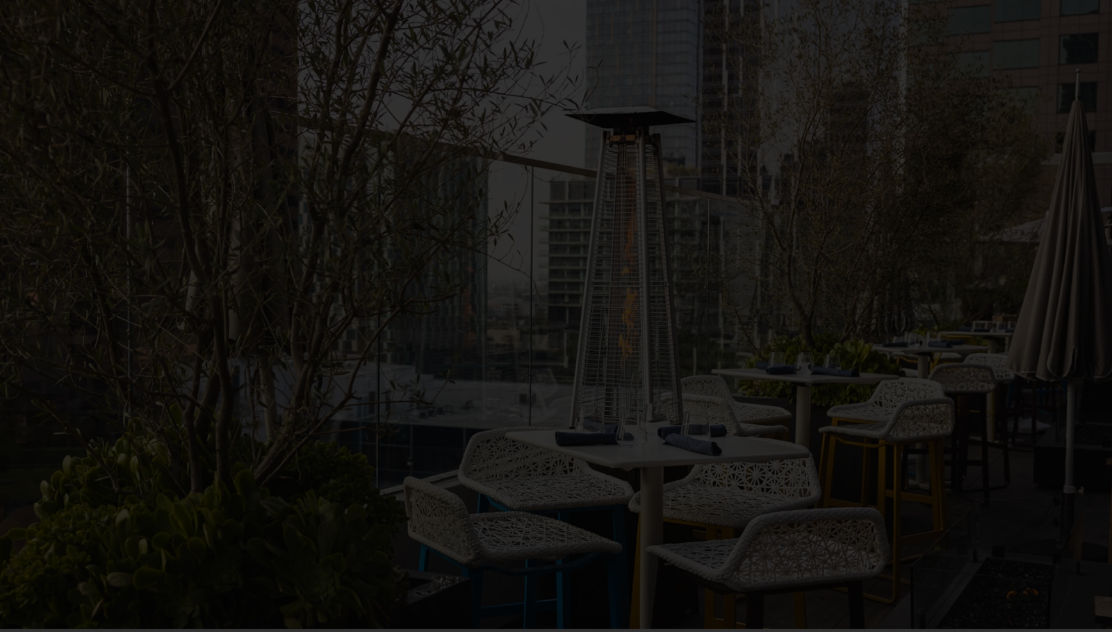 A dim outdoor patio with white chairs and tables, a tall heater, potted plants, and urban buildings in the background.