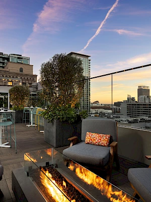 Rooftop lounge with fire pit, stylish gray seating, orange cushions, potted trees, bar area, and city skyline at sunset. Ends with a period.