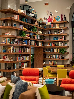 A cozy, modern library lounge with wooden bookshelves, colorful chairs, plants, and quirky decor—perfect for reading and socializing.