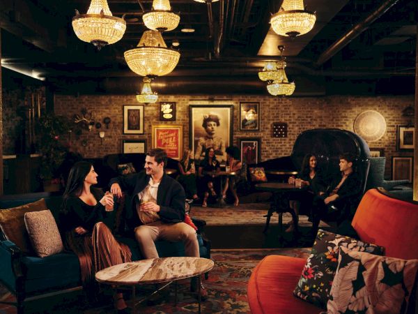 A cozy, dimly lit lounge with people chatting on sofas and chairs, vintage decor, chandeliers, and framed art on warm-toned walls, relaxed vibe.