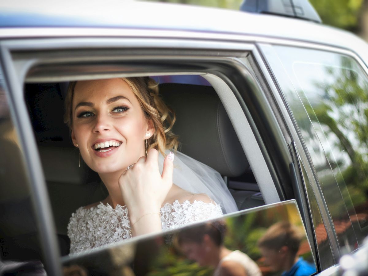 A smiling bride-to-be sits in a car window, adjusting her veil and posing for a quick photo inside the vehicle.