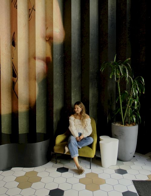 A woman sits in a yellow chair inside a modern lobby with tall striped panels, a potted plant, and geometric tiled floor.